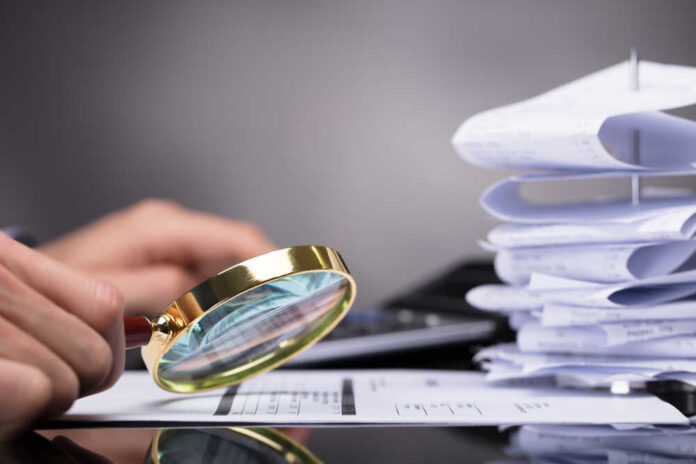 Close-up,Of,A,Businessperson's,Hand,Looking,At,Invoice,Through,Magnifying