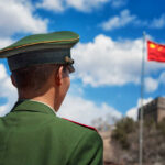 Chinese Cameras EXPOSE U.S. Vulnerability Soldier in a green military uniform facing the Great Wall of China with the Chinese flag in the background