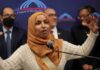 A woman in a hijab passionately speaking at a podium with a diverse group in the background