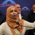 A woman in a hijab passionately speaking at a podium with a diverse group in the background