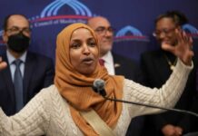 Omar EXPLODES During Trump Speech — Backfires Horribly A woman in a hijab passionately speaking at a podium with a diverse group in the background