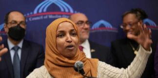 A woman in a hijab passionately speaking at a podium with a diverse group in the background