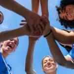 Voters React to Maine Democrat’s Sports Comments Group of female athletes joining hands in a circle under a blue sky