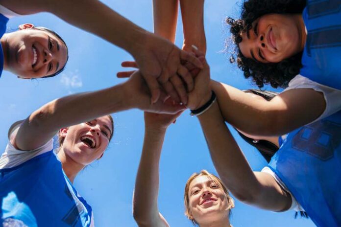 shutterstock_2704419661.jpg Group of female athletes joining hands in a circle under a blue sky