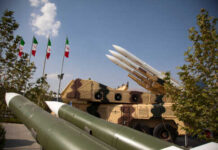 Ceasefire Whiplash: Iran Launches Missiles Military missiles displayed outdoors with Iranian flags in the background