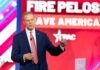 Nazi Reference by Historian Stirs Political Storm A man in a suit speaking at a political conference with a backdrop displaying campaign slogans