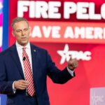 Nazi Reference by Historian Stirs Political Storm A man in a suit speaking at a political conference with a backdrop displaying campaign slogans