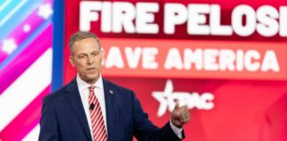 A man in a suit speaking at a political conference with a backdrop displaying campaign slogans