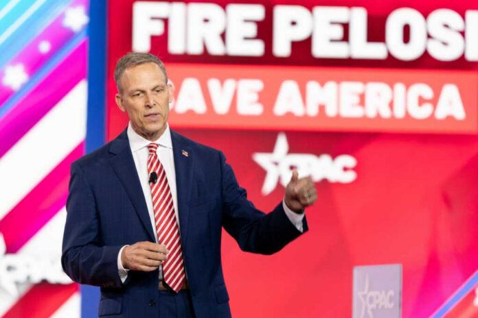 A man in a suit speaking at a political conference with a backdrop displaying campaign slogans