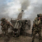 Soldiers fire a field howitzer as smoke and dust rise around them