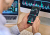 Shock Charges! Wall Street Icon Accused of Double-Dealing Person checking stock market graphs on phone and monitors.