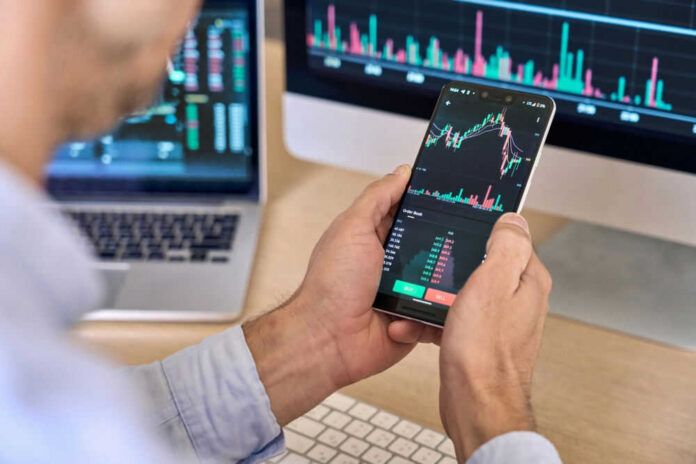 Person checking stock market graphs on phone and monitors.