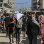Crowd walking through a dusty street between damaged buildings