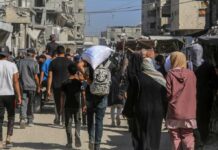 Crowd walking through a dusty street between damaged buildings