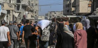 Crowd walking through a dusty street between damaged buildings