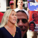 Greene’s Explosive Trump Accusation: Election Delay? Woman speaking at outdoor event with protest signs in background