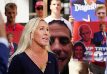 Greene’s Explosive Trump Accusation: Election Delay? Woman speaking at outdoor event with protest signs in background