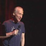 Media Spin Exposed? Maher Crashes Kirk Film Man holding microphone on stage in dark setting