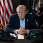Trump Puts Review Wall Around AI A serious-looking man seated at a table with phones and an American flag in the background