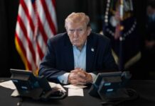 Trump Puts Review Wall Around AI A serious-looking man seated at a table with phones and an American flag in the background