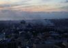 A city skyline at sunset with smoke rising from buildings