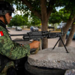 Soldier aiming machine gun on roadside under a tree.