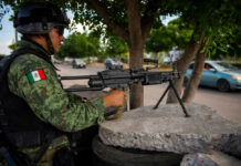 Soldier aiming machine gun on roadside under a tree.