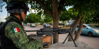 Soldier aiming machine gun on roadside under a tree.