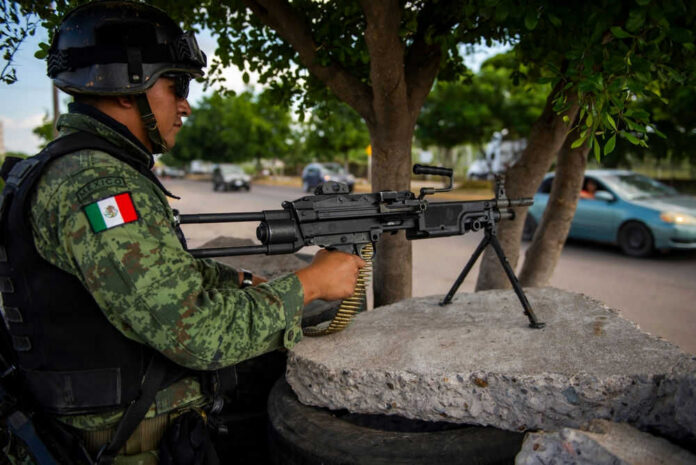 Soldier aiming machine gun on roadside under a tree.