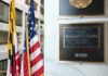 Office plaque beside U.S. and state flags in a government building