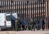 Border agents escort a group near a tall fence and a patrol van