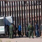 Meloni Threatens Schengen — Spain Fumes Border agents escort a group near a tall fence and a patrol van
