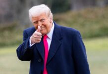 Trump’s Media War Turns Menacing A man in a dark coat and pink tie giving a thumbs up outdoors
