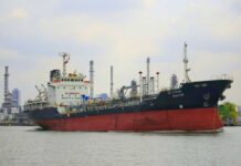 A cargo ship navigating through a port with industrial buildings in the background