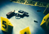 TDS Claim Hijacks Double Murder A handgun on a table with evidence markers and crime scene tape in the background