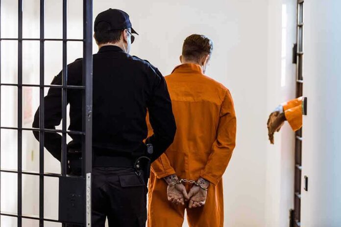shutterstock_1030254064-3 Prisoner in orange jumpsuit handcuffed and escorted by officer