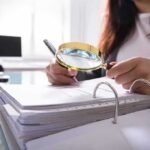 Person using a magnifying glass to review documents in a binder