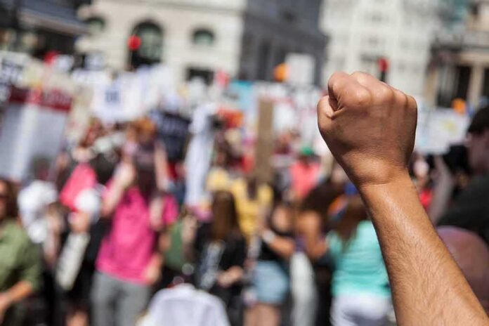 shutterstock_1135501646-3 Raised fist in foreground at a crowded street protest