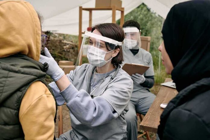 shutterstock_2042550827-1 Healthcare worker in protective gear examines a patient at an outdoor clinic