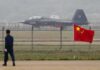 Stealth fighter jet on runway behind fence with Chinese flag in foreground