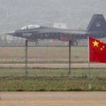 Stealth fighter jet on runway behind fence with Chinese flag in foreground