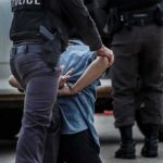 Police officer detaining a handcuffed person beside a vehicle