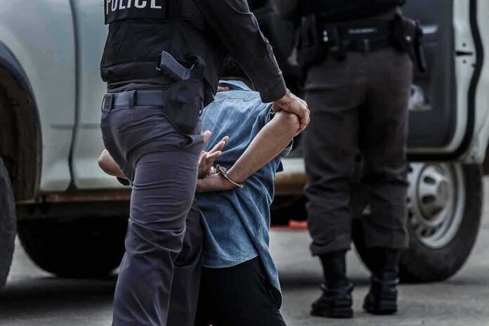 shutterstock_659470525-1 Police officer detaining a handcuffed person beside a vehicle