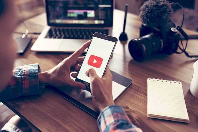 shutterstock_1041026248-10 Hands holding smartphone with video app icon at a workspace