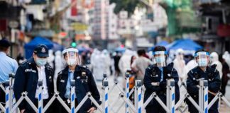 Police officers in masks and face shields stand behind a street barricade