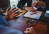 Crypto Godfather’s Wild Double Life Exposed Two people discuss documents at a desk with handcuffs in view