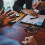 Crypto Godfather’s Wild Double Life Exposed Two people discuss documents at a desk with handcuffs in view