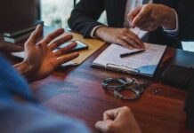 Crypto Godfather’s Wild Double Life Exposed Two people discuss documents at a desk with handcuffs in view