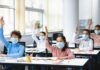 Schoolchildren wearing masks raise hands in a classroom