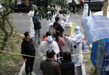Chemical Panic Triggers $125M Reckoning Christian Jones and others wait at an outdoor COVID-19 testing site with staff in protective suits